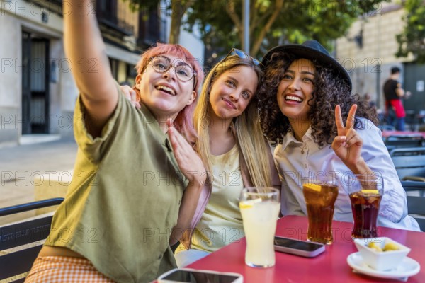 Happy latin and caucasian women taking selfie and gesturing success in an outdoor bar