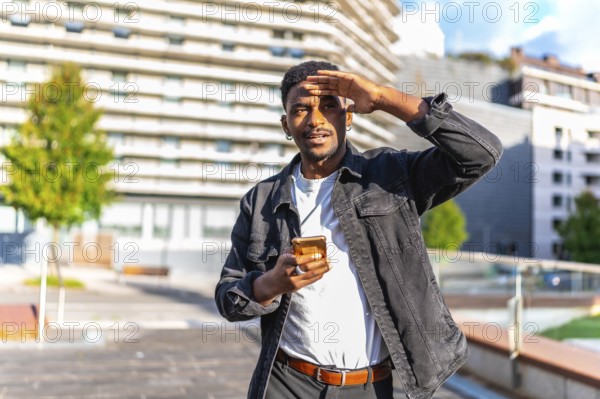 African man covering eyes from the sun while using mobile phone in the city searching for a location