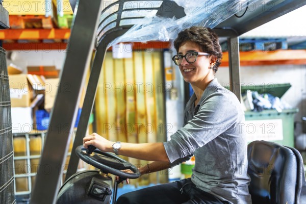Side view of a happy caucasian woman with short hair and eyeglasses driving forklift in a metallurgic factory