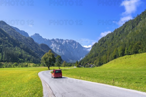 Red van driving along a winding road in the picturesque logarska dolina valley, surrounded by lush meadows, towering mountains, and a clear blue sky