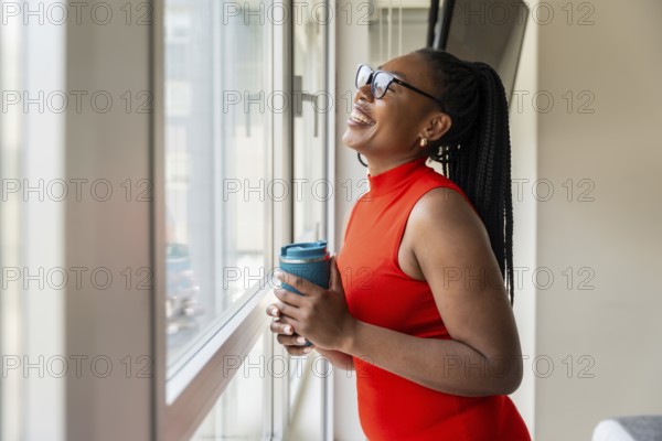 Happy businesswoman holding reusable coffee cup and looking out window during business trip, enjoying city view