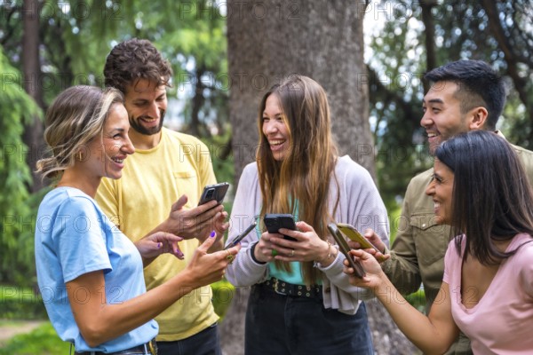 Smiling multiethnic group with phones watching internet or social networks in the park, technology concept