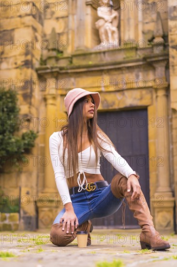 A young Latin woman with a white t-shirt, jeans and a hat enjoying the summer holidays, with a take away coffee, in a historic center, Pasaje San Juan, Gipuzkoa, Basque Country