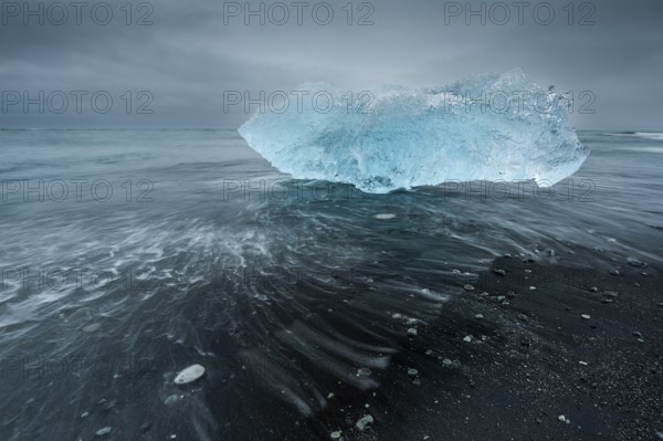 A shimmering blue iceberg is washed up by waves on a dark beach, Diamond Beach, Jökulsárlón glacier lagoon or Jökulsarlon, South Iceland, Iceland, Europe
