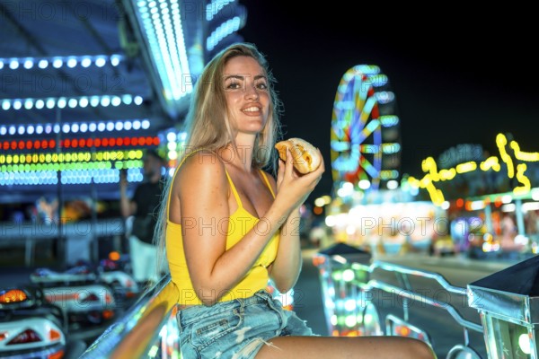 Blonde woman sitting eating a traditional hot dog at night fair