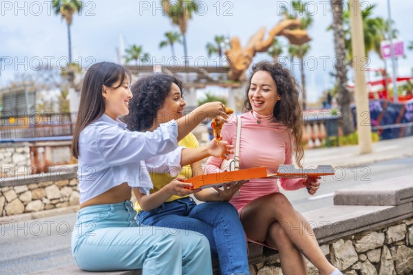 Three multi-ethnic female friends eating pizza sitting in a bench in the city