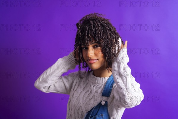 Young woman enjoying music with wireless headphones, wearing a white sweater and denim overalls, isolated on a vibrant purple background