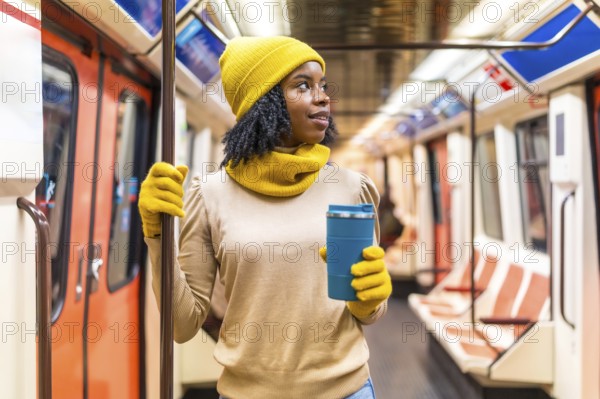 Young black woman wearing yellow winter clothes, holding a reusable coffee cup and traveling on a subway train
