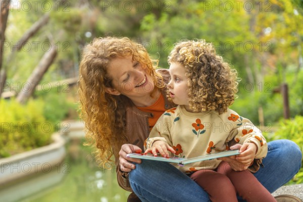 Cute girl and smiling caucasian mother reading a book sitting on a park together