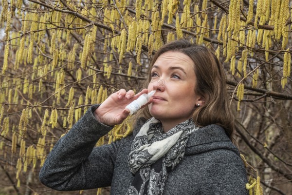 Young Caucasian woman with allergy problem stands by flowering hazel bush and sprays her nose in Ystad, Skåne County, Sweden, Scandinavia, Europe