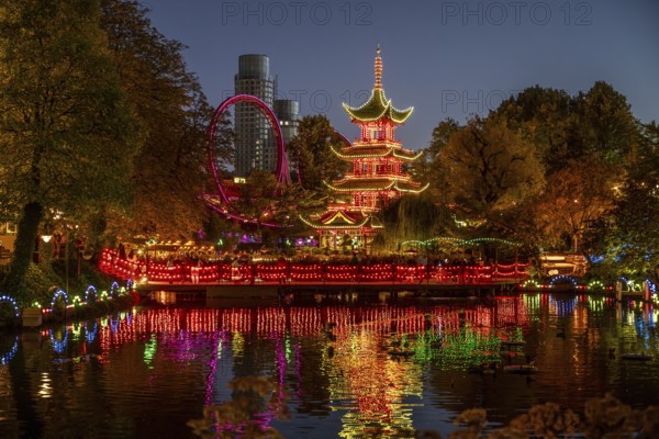 Illuminated pagoda, colourful lighting, roller coaster, trees, cityscape at night with modern building, Tivoli amusement and recreation park, Copenhagen, Denmark, Europe