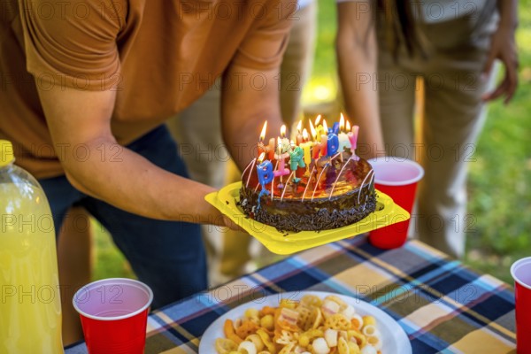 Close-up of the hands of a young man holding birthday cake with candles during picnic with friends