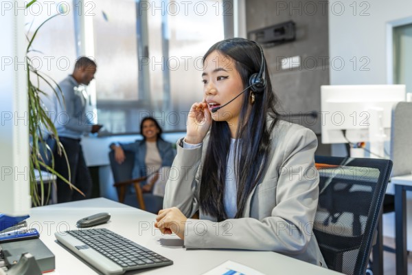 Young asian operator talking with headset and working on computer in a coworking office, with colleagues talking in the background