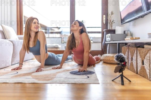 Two women recording an online yoga class from home, performing the cobra pose on a rug in the living room