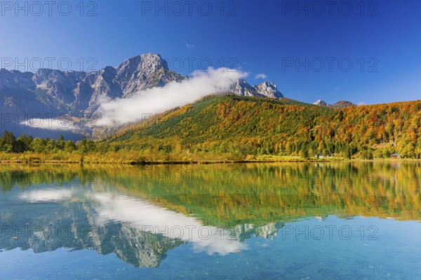 Lake Almsee in autumn with a forest reflected in the lake. A slowly dissipating fog moves over the lake. Blue sky with no clouds