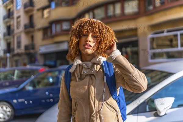 Cool young latin woman with curly hair and casual clothes walking carrying rucksack along a city next to cars and buildings
