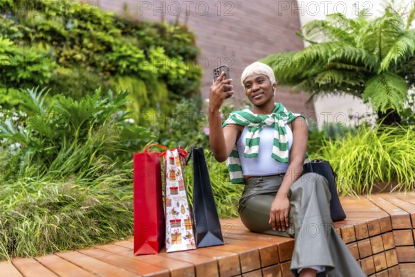 African woman taking selfie sitting next to colorful shopping bags outside a mall after shopping