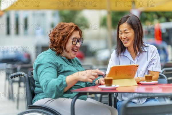 Woman with disability using tablet next to an asian female friend sitting in an outdoor cafeteria