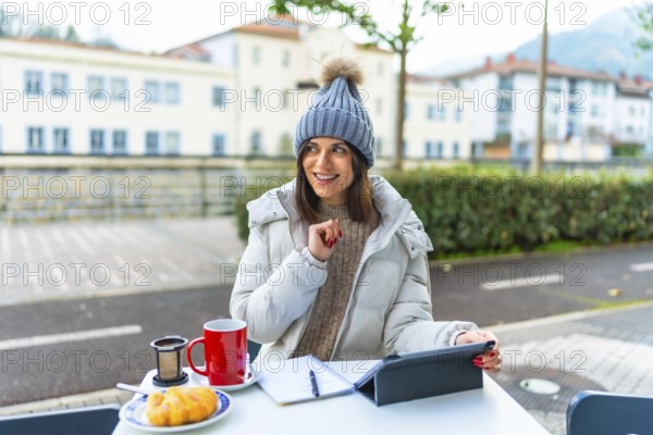 Creative caucasian beauty woman with warm clothes using digital tablet while drinking tea sitting on an outdoor cafeteria