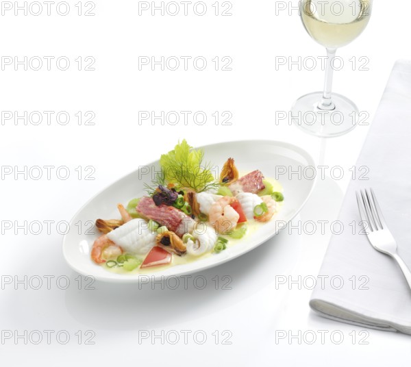 A plate with a fish dish, a glass with white wine, a fork on a napkin, white tablecloth, studio shot