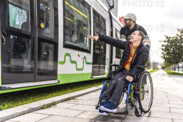 Young man in a wheelchair points at an approaching tram while his friend stands behind him, offering support and companionship