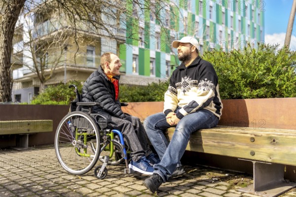 Two men, one in a wheelchair, enjoy a friendly conversation on a park bench in a modern urban setting
