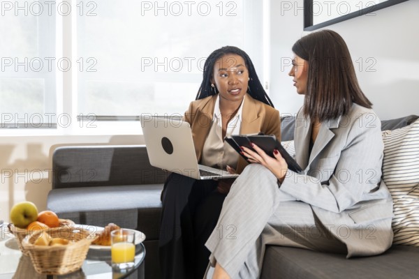 Two businesswomen using laptop and tablet, discussing work during business trip in hotel room