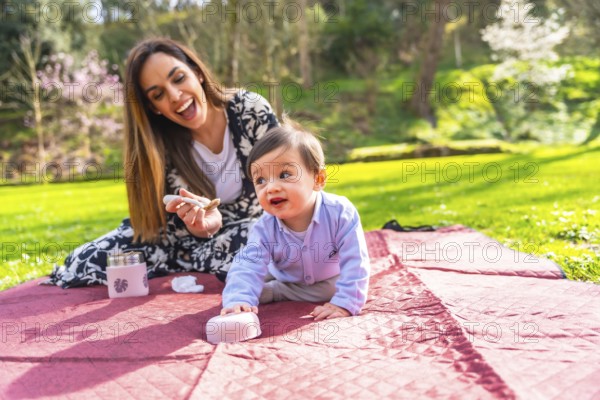 Mother giving food to her baby son during a picnic on a blanket in a park, surrounded by green grass and trees