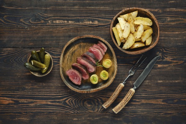 A beautifully arranged dinner features thinly sliced meat alongside roasted potatoes and pickles. The rustic wooden table enhances the warm, inviting atmosphere of the meal
