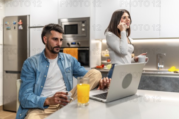 Young couple working from home in the kitchen, having breakfast and using laptop and smartphone