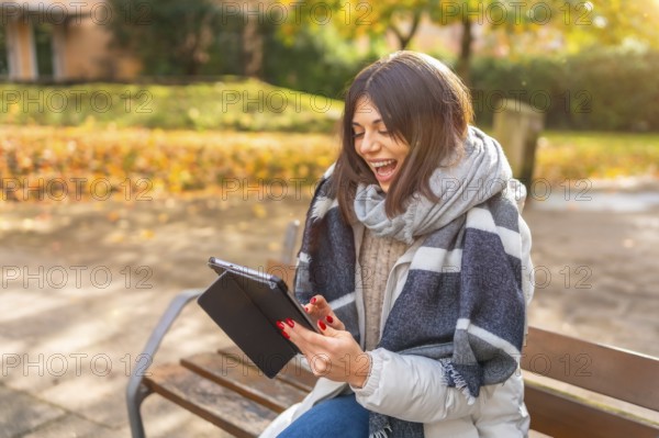 Rays of the sun illuminating a caucasian happy woman laughing while using digital tablet in a park in a cold day of autumn