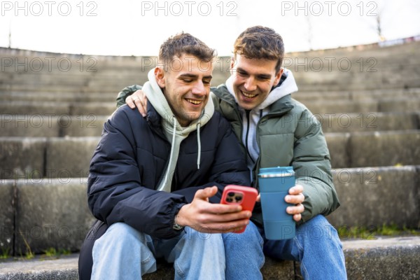 Two men are sitting on steps, embracing and looking at a smartphone while one holds a reusable coffee cup, enjoying a moment of togetherness