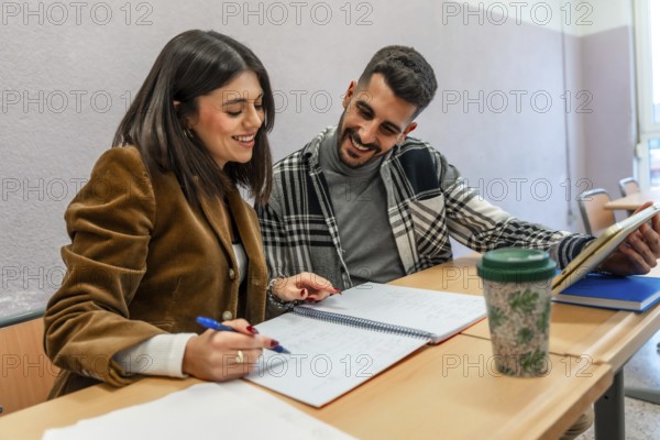 Two happy university students are studying together in a classroom, taking notes and using a tablet, enjoying the learning process