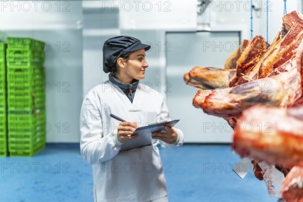 Woman in uniform and hat writing on clipboard checking goods in a meat processing factory