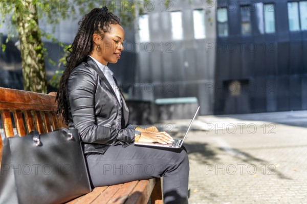 Latin beauty and elegant businesswoman working on remote using laptop sitting on a bench sunbathing in the financial district