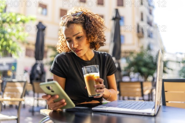 Young arab woman drinking iced coffee and using mobile phone while working with laptop in an outdoors cafeteria