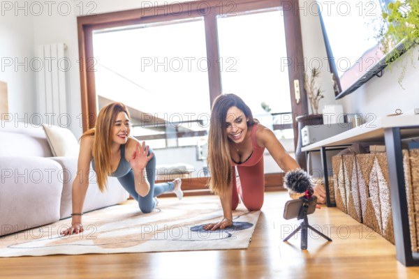 Two women in sportswear smiling and recording an online yoga class in their modern living room, embracing wellness and connection
