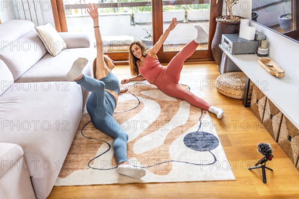Two smiling women lying on carpet practicing yoga together at home following online class on television