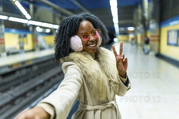 Smiling woman wearing earmuffs and pink glasses making selfie and victory sign while waiting subway train in station platform