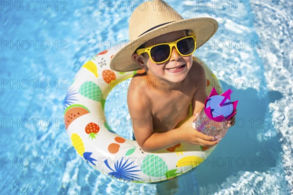 Top view close-up of a funny caucasian boy inside a pool drinking water into a rubber ring colorful float smiling at camera