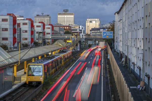 Essen, North Rhine-Westphalia, Germany, motorway A40 in the city centre, exit Essen Zentrum, during dusk. Underground line U18 on the way to Berliner Platz stops at Savignystrasse underground station. On the right, residential buildings behind a noise barrier. A LANUV air monitoring station, Essen Kruppstrasse 117 (EKRU2), is located on the wall on the right under the lantern. Residents are affected by noise and exhaust fumes. Building with Postbank, Eon and Evonik lettering at the back, Europe