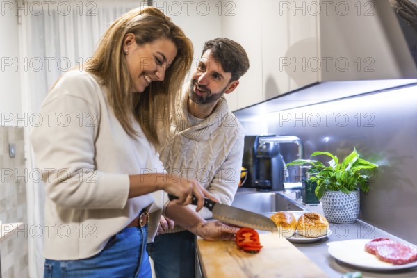 Cheerful couple making hamburgers together, slicing tomatoes and having fun in their modern kitchen