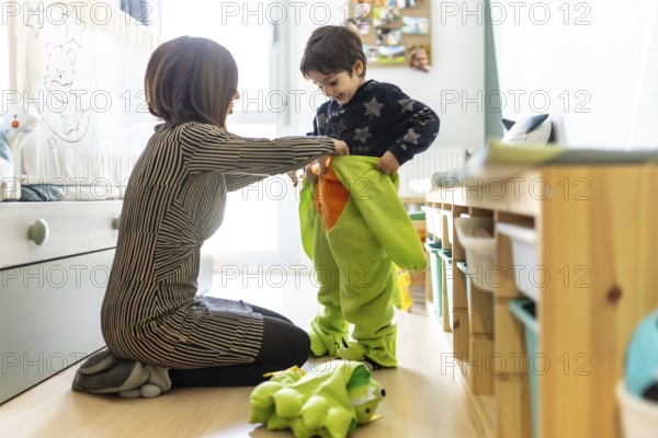 Mother assisting her son in putting on a colorful dinosaur costume, enjoying playful moments together in the cozy bedroom