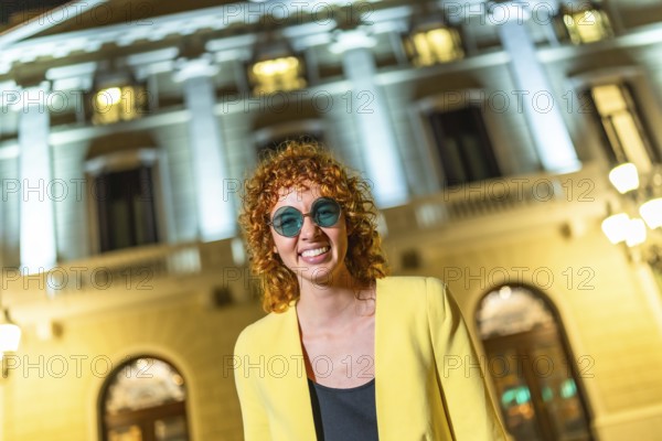 Modern woman in yellow casual clothes and sunglasses laughing at camera standing in the city at night