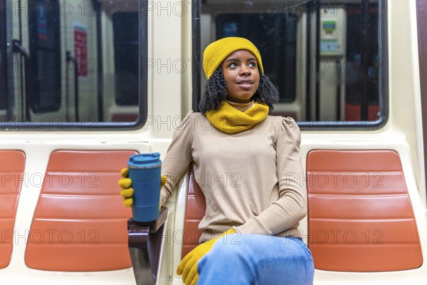 Young black woman wearing yellow winter clothes holding reusable coffee cup while riding subway train