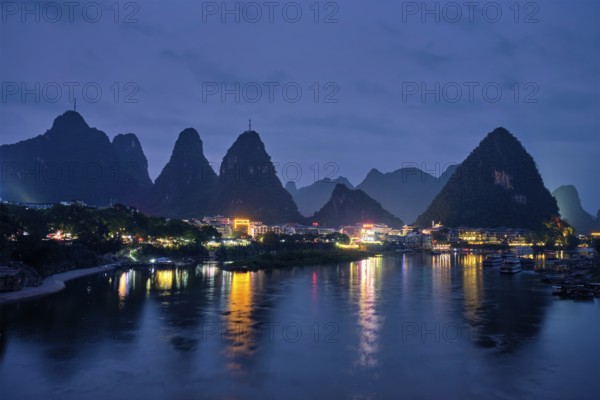 View of Yangshuo town illuminated in the evening with dramatic karst mountain landscape in background over Li river. Yangshuo, China, Asia
