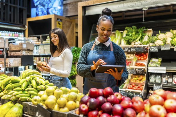 Friendly saleswoman using digital tablet and managing organic products while customer choosing fresh fruits in supermarket