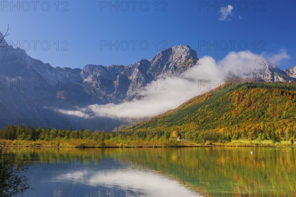 Lake Almsee in autumn with a forest reflected in the lake. A slowly dissipating fog moves over the lake. Blue sky with no clouds