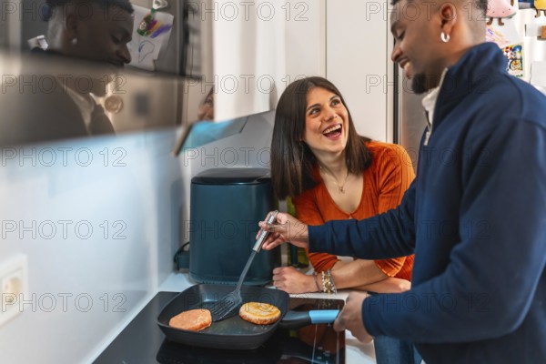 Cheerful multiethnic couple laughing and having fun while preparing burgers together on the stovetop in their modern kitchen