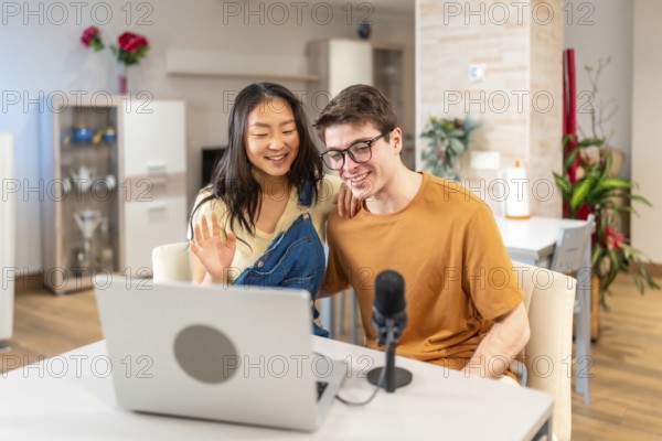 Happy multiethnic couple waving at the laptop camera while recording a vlog or streaming a podcast from home